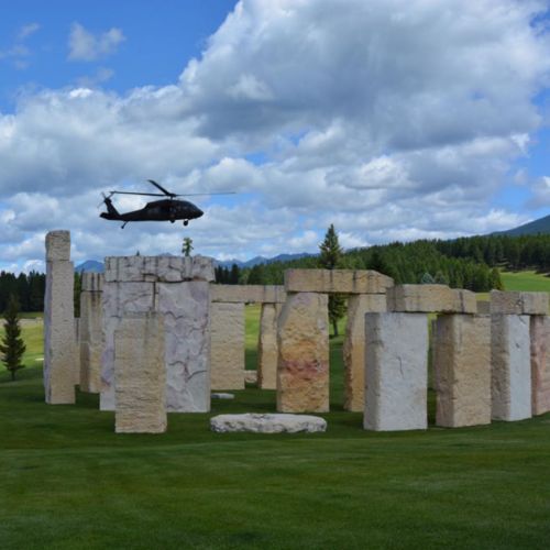 A helicopter flies over a stone structure resembling Stonehenge, set in a grassy landscape under a cloudy sky.