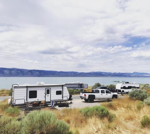 Two RVs parked near a lake, surrounded by dry grass and shrubs, with mountains in the background under a cloudy sky.