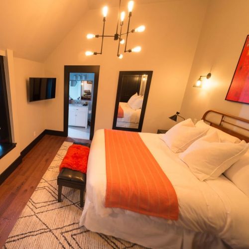Cozy bedroom with a large bed, orange accents, modern lighting, and a TV, featuring a stylish rug and mirrors.