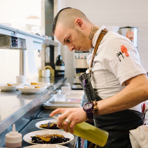 A chef in a kitchen carefully drizzles sauce over plated dishes, focused on presentation.