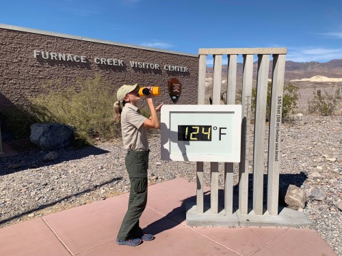 A park ranger at Furnace Creek Visitor Center checks a thermometer displaying 124°F in a desert landscape.