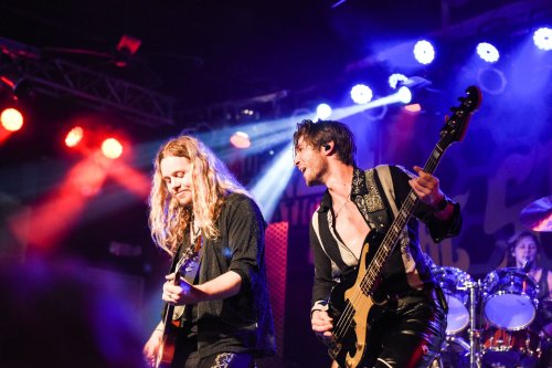 Two musicians perform on stage, one playing guitar and the other on bass, with colorful lights and a drummer in the background.