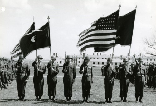 Soldiers in uniform holding flags during a military ceremony, with a formation of troops in the background.