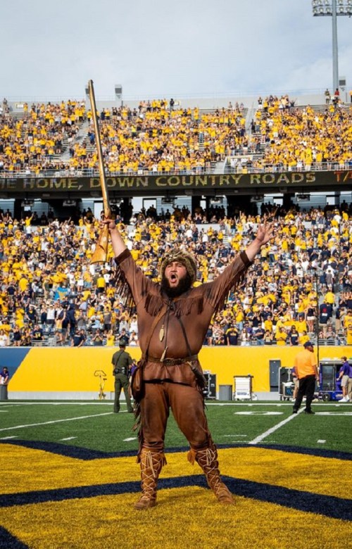 A costumed mascot in brown attire raises a rifle, cheering in front of a packed stadium filled with fans in yellow.
