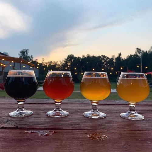 Four glasses of craft beer in varying colors, set against a sunset backdrop with string lights in the distance.