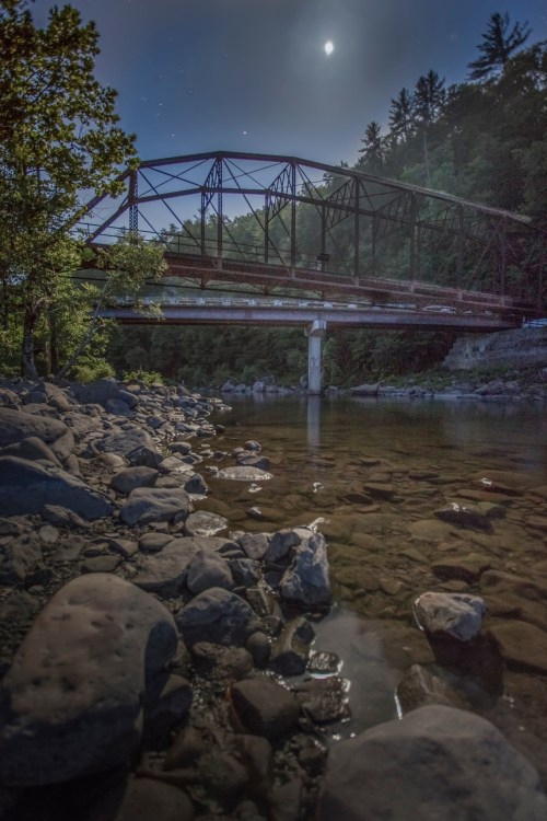 A moonlit scene of a rustic bridge over a calm river, surrounded by trees and rocky riverbank.