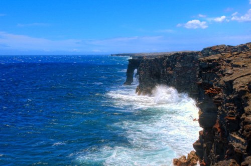 Waves crash against rugged cliffs under a bright blue sky, creating a scenic coastal view.