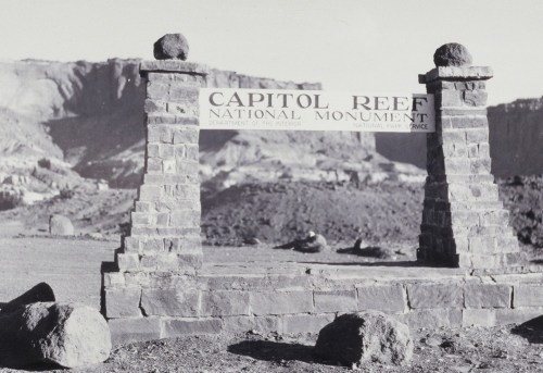 Black and white photo of the Capitol Reef National Monument sign, framed by stone pillars and rocky landscape.
