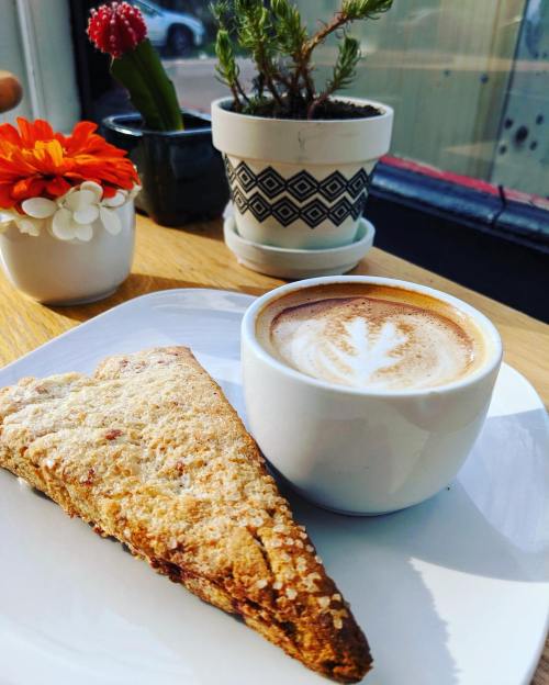 A slice of scone on a plate next to a latte with a leaf design, set on a wooden table with plants in the background.