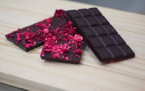 Three pieces of dark chocolate, one topped with crushed raspberries, on a wooden surface.