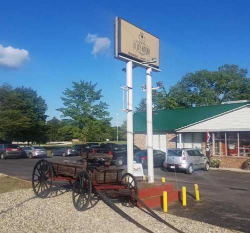 A rustic wagon in front of a restaurant with a sign, surrounded by trees and parked cars under a clear blue sky.