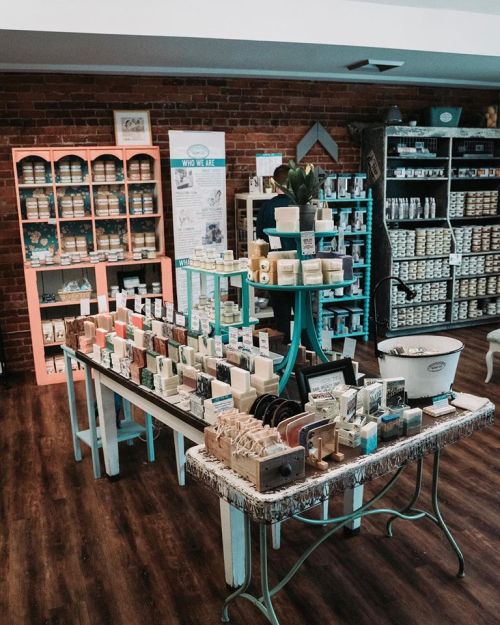 A cozy shop interior featuring a display of colorful soaps on tables and shelves against a brick wall.