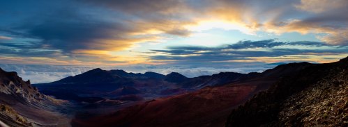 Panoramic view of a mountainous landscape at sunset, with clouds below and a colorful sky above.