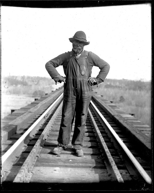 A man in overalls and a hat stands confidently on a railway bridge, hands on hips, with a landscape in the background.