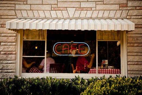 A cozy café window with a neon "CAFE" sign, showcasing diners at checkered tables inside.