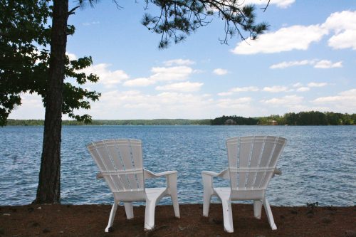Two white Adirondack chairs facing a serene lake under a blue sky with fluffy clouds and trees in the background.