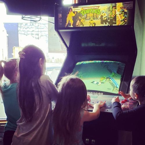 Four children playing an arcade game together, focused on the screen, with colorful graphics above them.