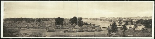 A panoramic view of a military camp with numerous tents and buildings, surrounded by trees and open fields.