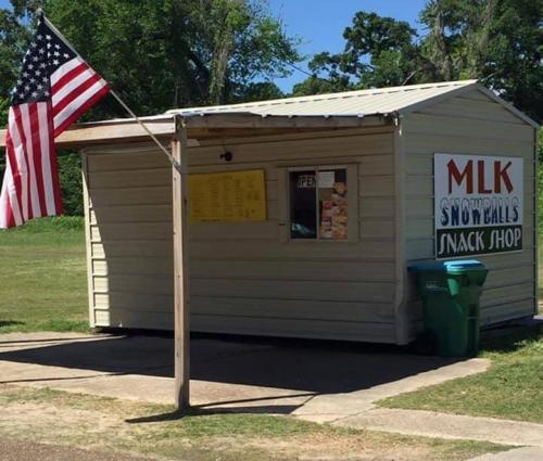 A small snack shop with a sign for "MLK Snowballs" and an American flag outside, set in a grassy area.
