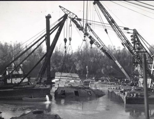 Black and white photo of cranes lifting a sunken boat from a river, with workers and equipment visible.
