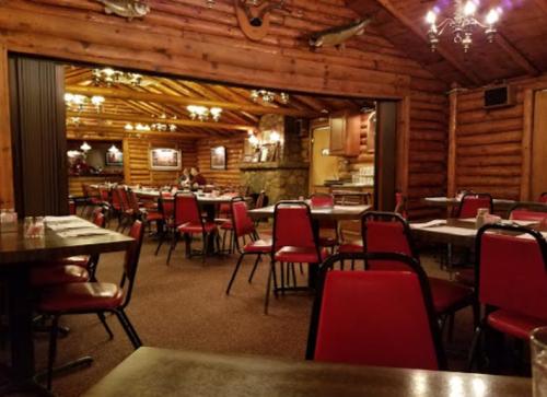 Cozy log cabin restaurant interior with wooden walls, red chairs, and tables set for dining. Warm lighting creates a welcoming atmosphere.