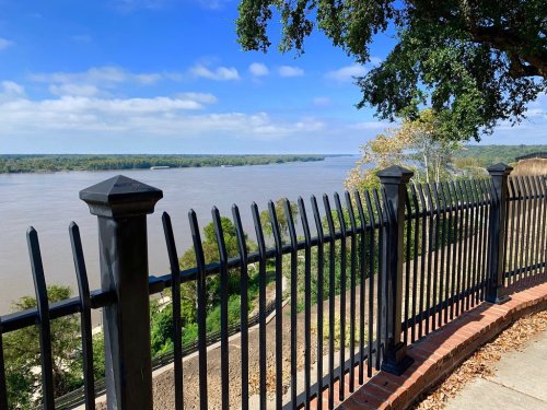 A scenic view of a river from a park, with a black fence and trees under a blue sky with scattered clouds.