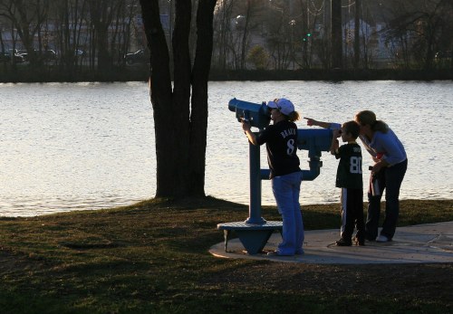 A woman and two children use a telescope by a lake during sunset, with trees in the background.