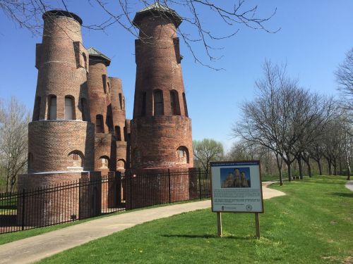 Historic brick towers stand beside a pathway, with a sign detailing their history, surrounded by trees and blue sky.
