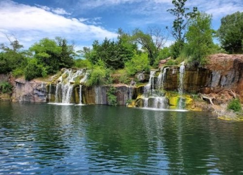 A serene waterfall cascading over rocky cliffs into a calm, greenish-blue lake surrounded by lush trees.