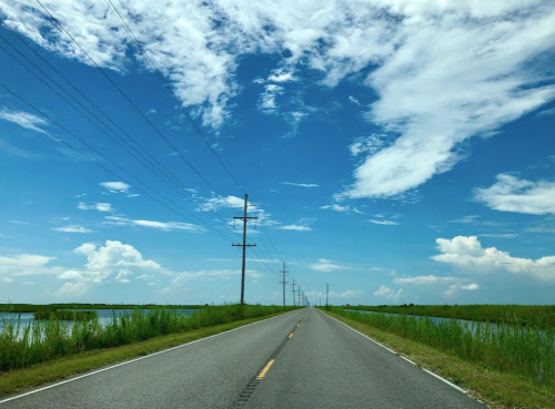 A straight road lined with power lines, flanked by water and lush greenery under a bright blue sky with scattered clouds.