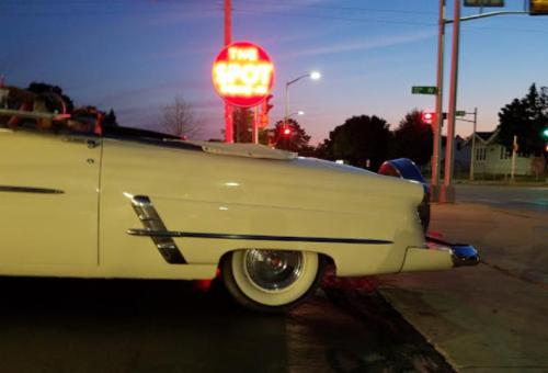A classic white convertible parked near a neon sign reading "The Spot" at dusk.