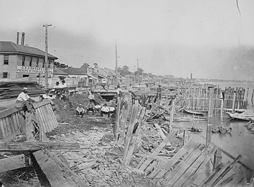 Historic waterfront scene with wooden structures, people sitting, and boats along the shore, showcasing early 20th-century life.