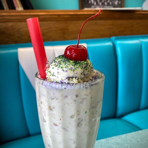 A creamy milkshake topped with whipped cream, a cherry, and colorful sprinkles, served in a glass with a red straw.