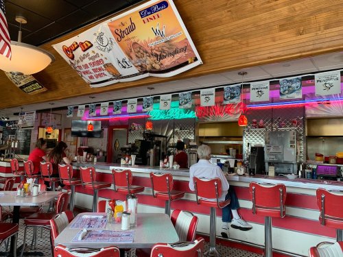A retro diner interior with red and white decor, bar stools, and a counter, featuring colorful banners and neon lights.
