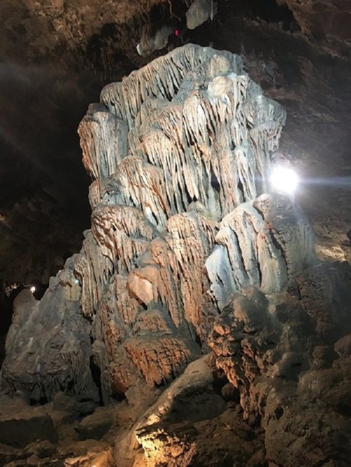 A large, textured rock formation in a cave, illuminated by lights, showcasing stalactites and stalagmites.