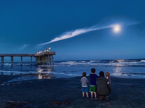 Four children and an adult stand on a beach at dusk, gazing at the moon and a pier illuminated by lights.