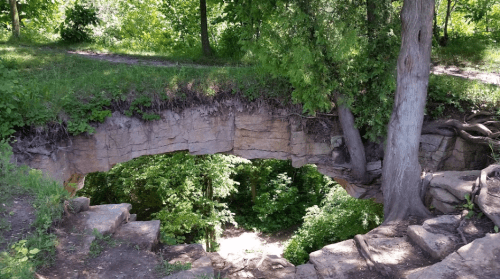 A natural stone arch surrounded by lush greenery and trees, with a path visible in the background.