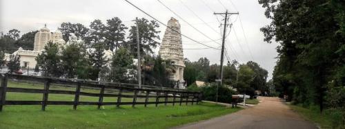 A view of a temple with intricate architecture, surrounded by trees and a rural road, under a cloudy sky.