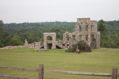 Ruins of a stone building surrounded by greenery, with a grassy area and a wooden fence in the foreground.