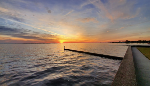 A serene sunset over a calm lake, with vibrant colors reflecting on the water and a wooden post extending into the scene.
