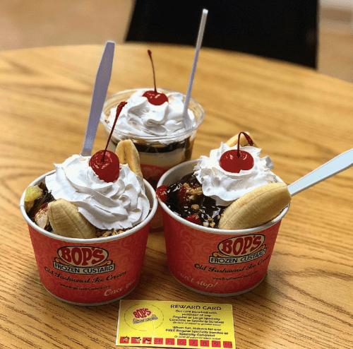 Three cups of frozen custard topped with whipped cream, cherries, and banana slices, on a wooden table with a reward card.
