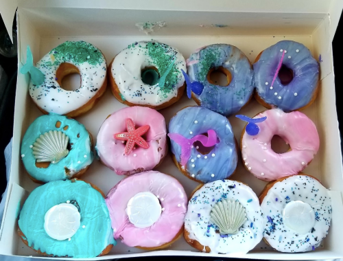 A box of colorful donuts decorated with ocean-themed icing, sprinkles, and edible decorations like shells and starfish.
