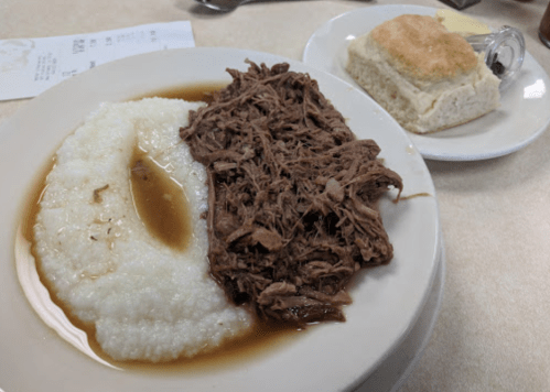 A plate with pulled beef, creamy grits, and a biscuit on the side, served with a brown gravy.
