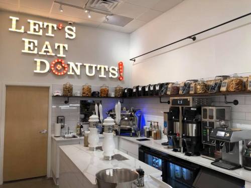 A bright café interior featuring a "Let's Eat Donuts!" sign, coffee machines, and jars of toppings on shelves.