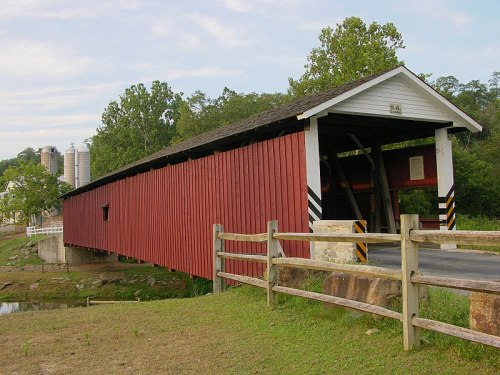 A red covered bridge spans a small stream, surrounded by greenery and a wooden fence. Silos are visible in the background.