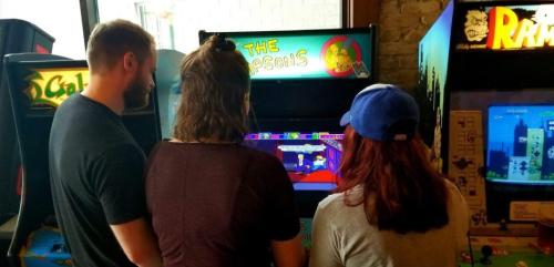Three people playing an arcade game, "The Simpsons," in a retro gaming environment.