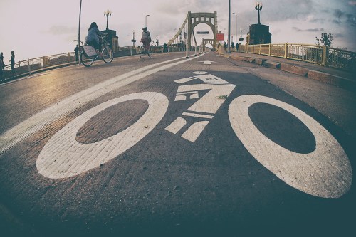 A low-angle view of a bike lane on a bridge, with cyclists in the background and a cloudy sky above.