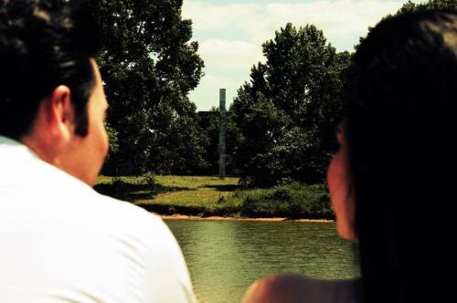 A couple sits by a lake, gazing at a distant landscape with trees and a building in the background.