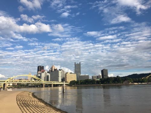 A view of Pittsburgh's skyline with a river in the foreground and a bridge, under a partly cloudy sky.