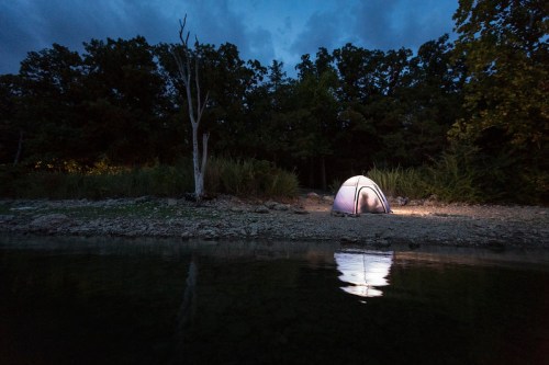 A lit tent by a calm water body at dusk, surrounded by trees and a darkening sky.